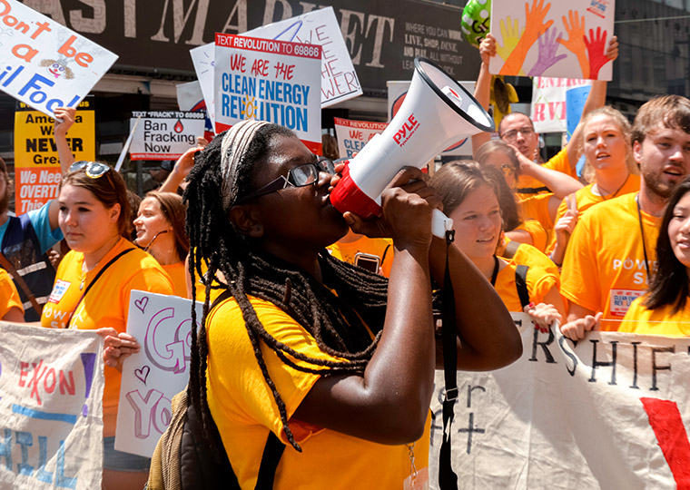 In the forground, a young person speaks into a bullhorn, with marchers in organge t-shirts and "clean energy revolution" signs can be seen behind them.