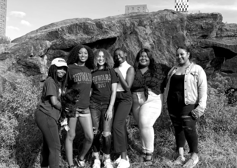 A black and white photo of 6 people smiling and standing side by side in front of a big rock.