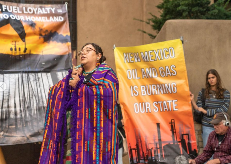 A person is holding a microphone in mid-speech. They're wearing glasses, dangling earrings, and a multi-colored shawl. The sign behind them says, "New Mexico Oil And Gas Is Burning Our State"