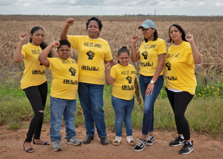 2 young children and 4 other people stand next to each other with their fists up in the air. They are all wearing yellow shirts that say "Rise St. James" with a black first underneath. They are standing in front of a field