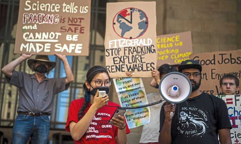People holding up brown cardboard signs. One reads "Science tells us FRACKING is NOT Safe and CANNOT be made safe". Someone stands with a megaphone speaker which is attached to a microphone someone with a red shirt holds up to their mouth. They're wearing black glasses and have a long side braid.