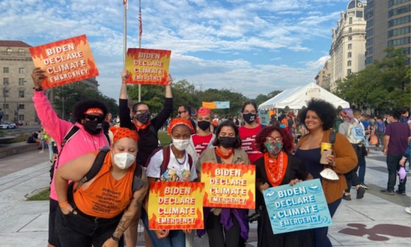PSN staff are smiling while holding up orange and yellow signs that read, "Biden declare climate emergency"