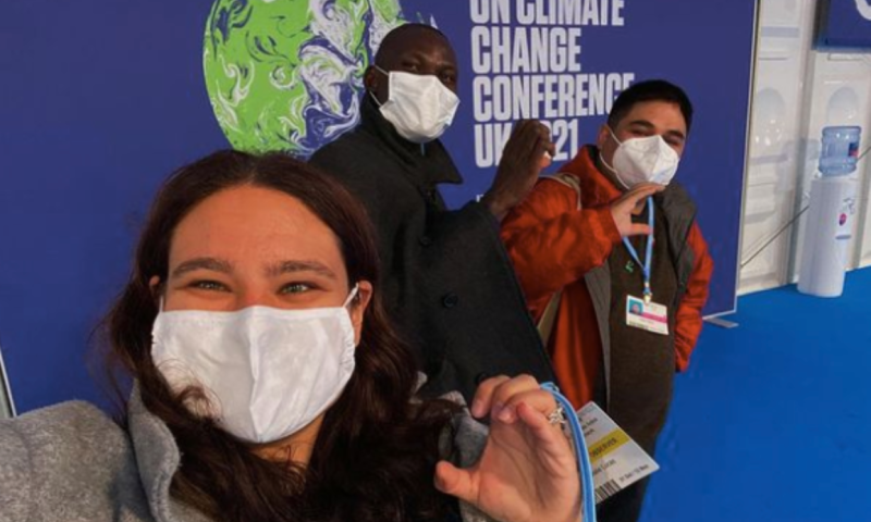 Three people wearing white masks covering their mask make a C shape with their hands. They are standing in front of a sign that reads "UN Climate Change Conference"