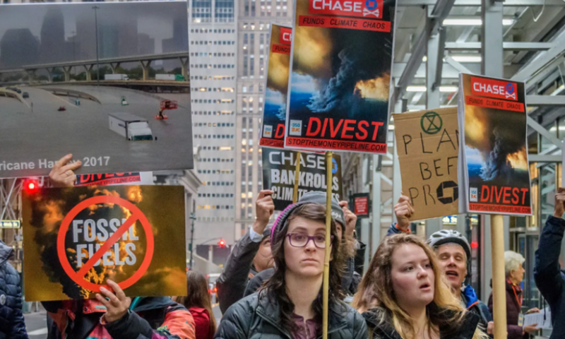 A group of people stand outside wearing jackets and carrying signs that read "Chase Funds Climate Chaos"