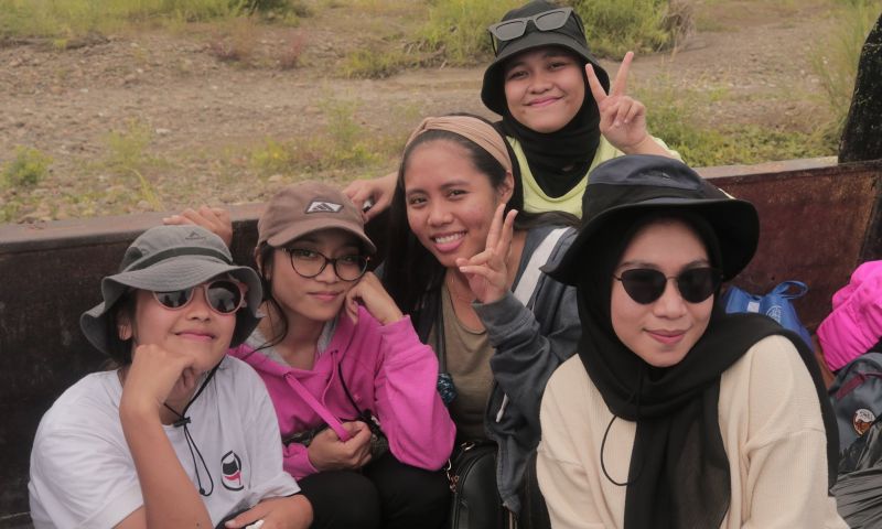 5 people are smiling at the camera. Two of them are holding up piece signs with their index and middle fingers. The person in the front has a black bucket hat on, black sunglasses, and a black scarf draped around their neck