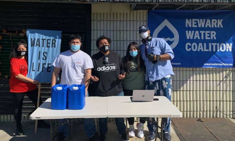 A person wearing a red shirt holds up a blue cloth sign that reads, "Water is life" while standing next to 4 other people standing in front of a gray table